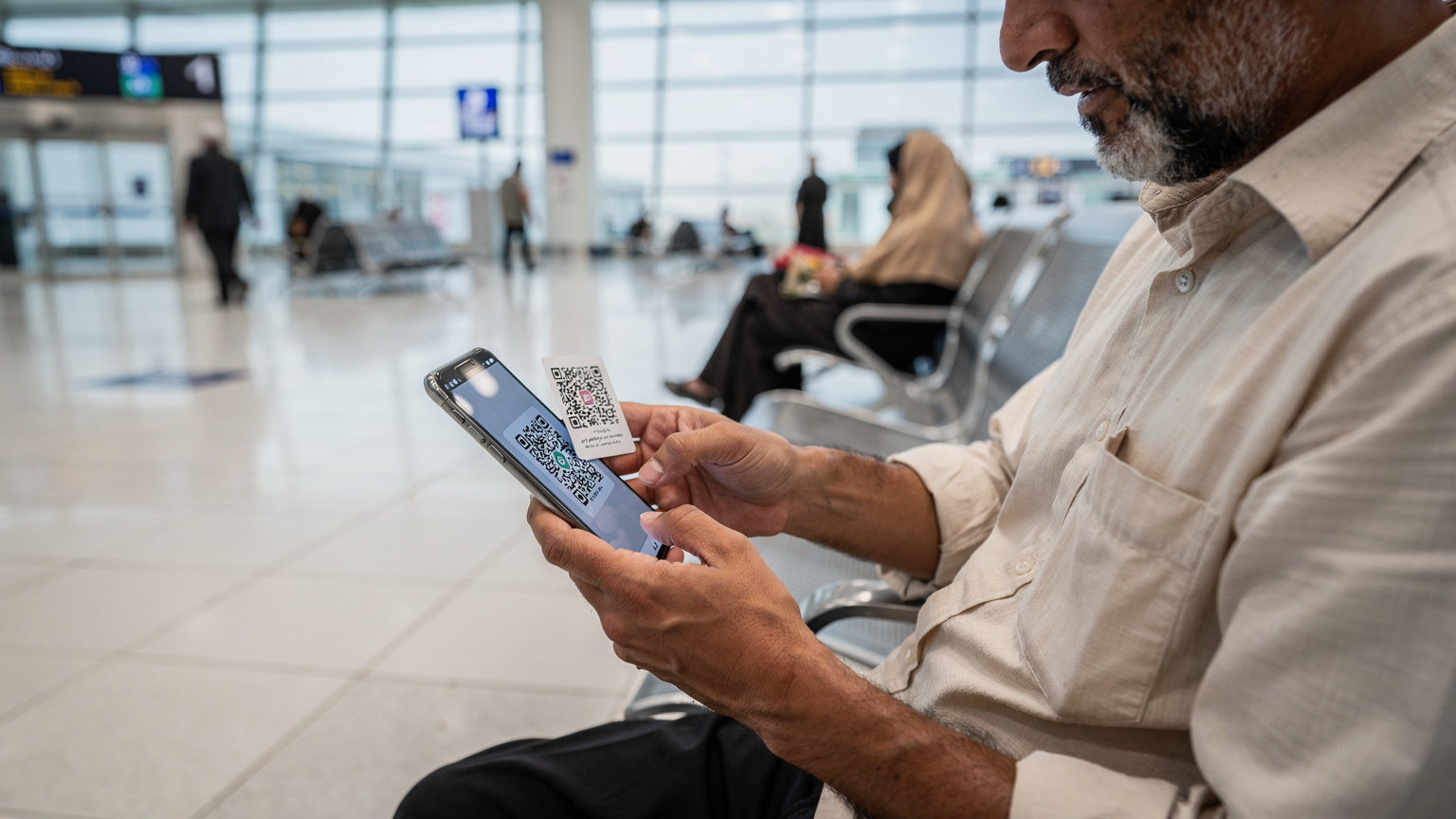 یک زائر میانسال با ریش در حال اسکن QR کد eSIM بر روی گوشی هوشمند خود در سالن انتظار فرودگاه بینالمللی نجف.