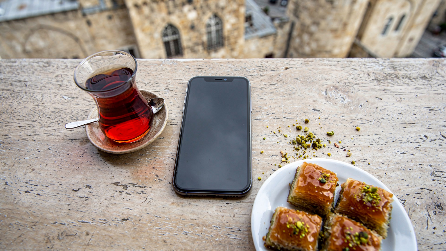 منظر علوي يظهر هاتفًا ذكيًا معروضًا على طاولة مقهى في الهواء الطلق في باكو، أذربيجان، مع فنجان من الشاي وأصابع بقلاوة، وخلفية ضبابية تظهر مباني المدينة القديمة.