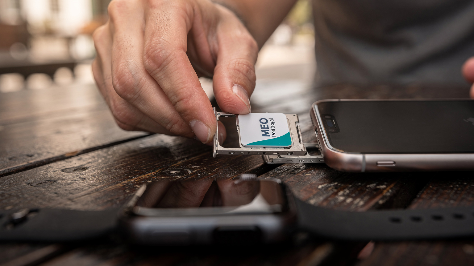 Eine Hand legt eine portugiesische MEO SIM-Karte in ein iPhone ein, während eine Apple Watch im Vordergrund leicht unscharf auf einem Holztisch liegt.