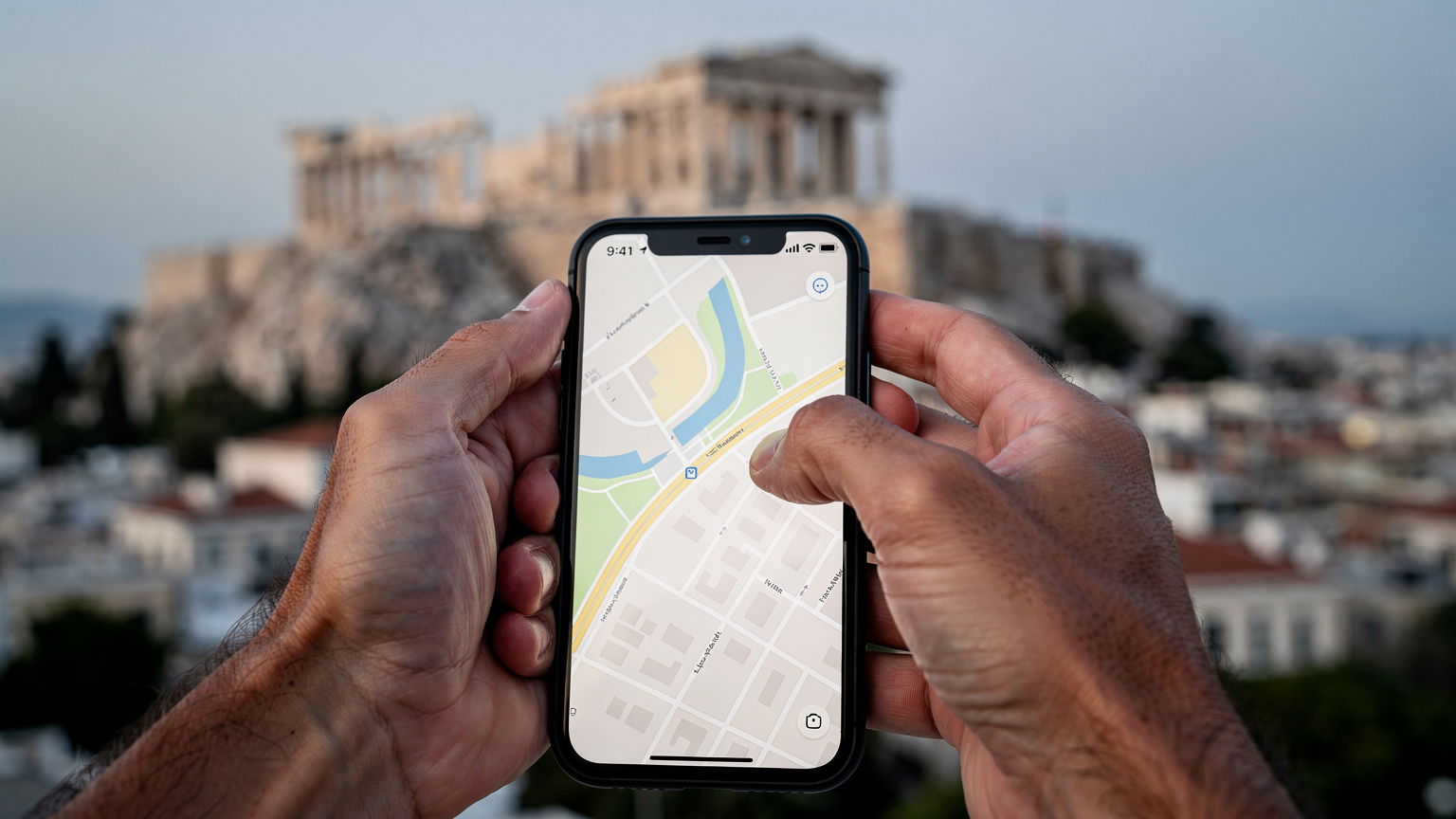 Närbild på ett par händer som håller en smartphone och bläddrar i en karta över Aten, med en bakgrund av Akropolis i diset.