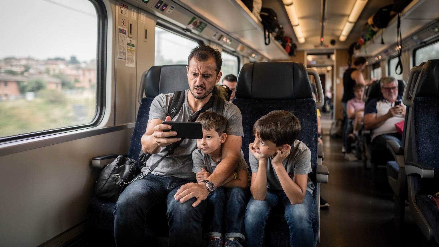 Un padre preoccupato tiene uno smartphone spento, mentre i suoi figli appaiono impazienti in un treno affollato, suggerendo una perdita di connettività.