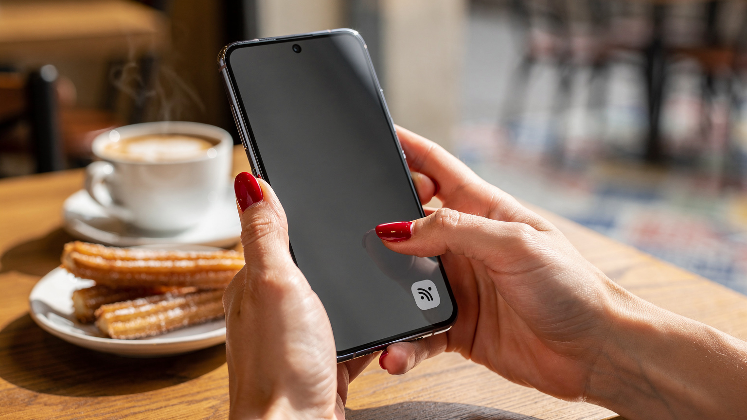 Manos de una mujer con las uñas pintadas de rojo, presionando con el pulgar la pantalla de un Samsung Galaxy S25 para activar una configuración de red, con una taza de café a un lado.