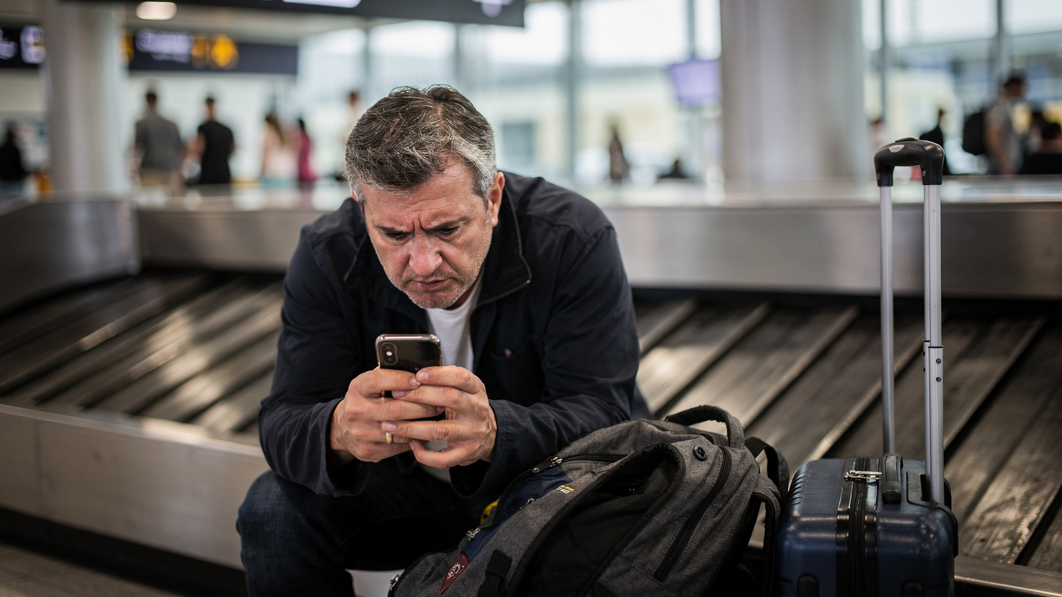 Un hombre con expresión de frustración mira su teléfono móvil mientras espera en la zona de recogida de equipajes del aeropuerto de Barcelona-El Prat, con sus maletas a un lado.
