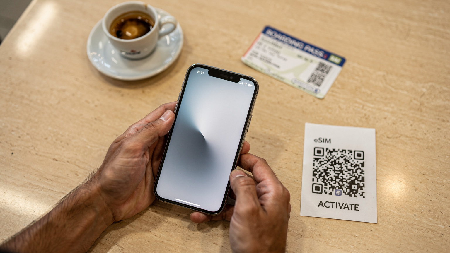 Mãos de um viajante segurando um iPhone 17, com o menu de configurações do eSIM aberto e um código QR de ativação em um papel ao lado, em um café tranquilo no aeroporto antes do embarque.