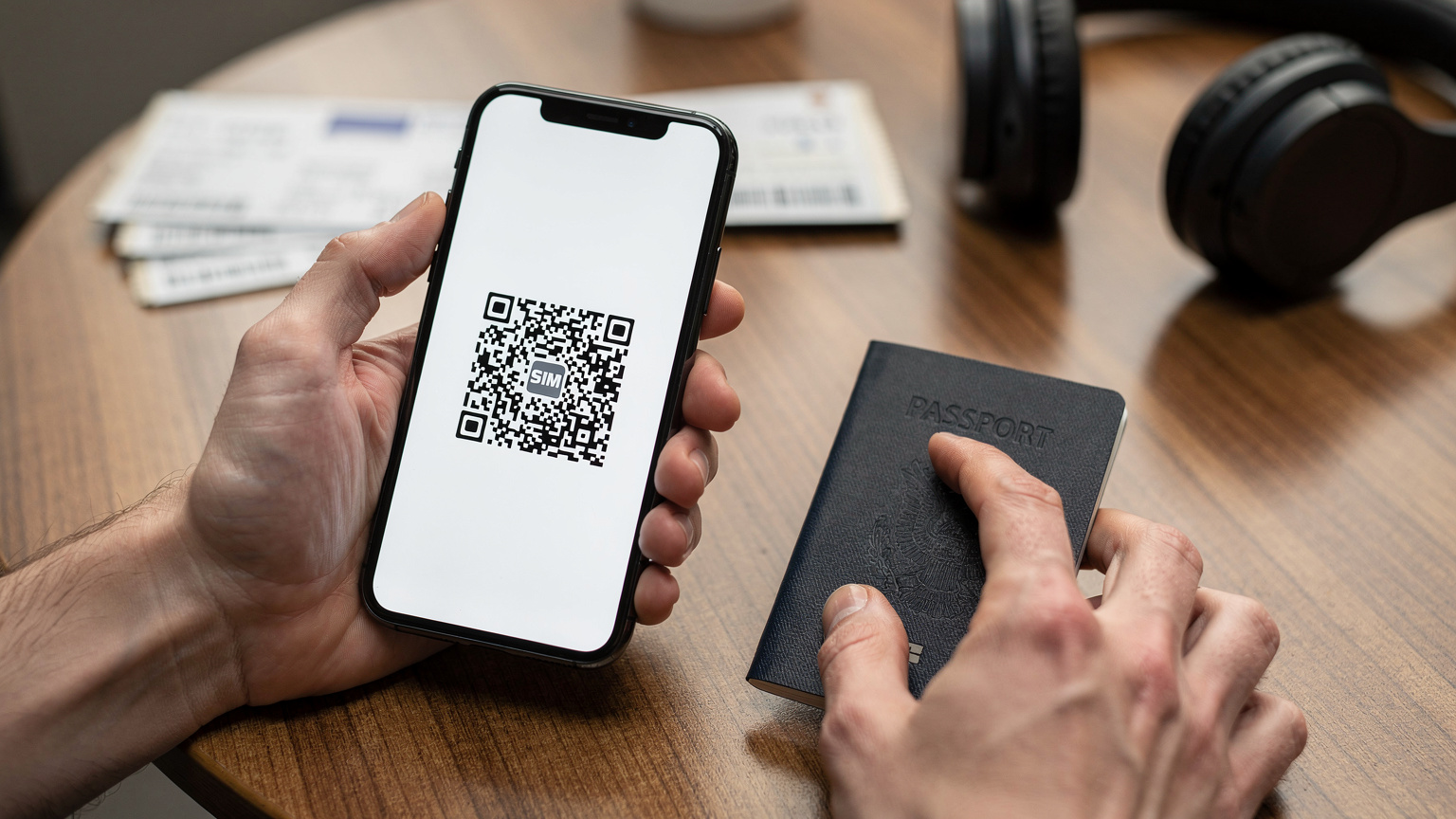 Детайлен кадър на ръце, които държат смартфон, показващ приложение за eSIM активация с QR код, до паспорт и билети за полет на масата в чакалня на летище.