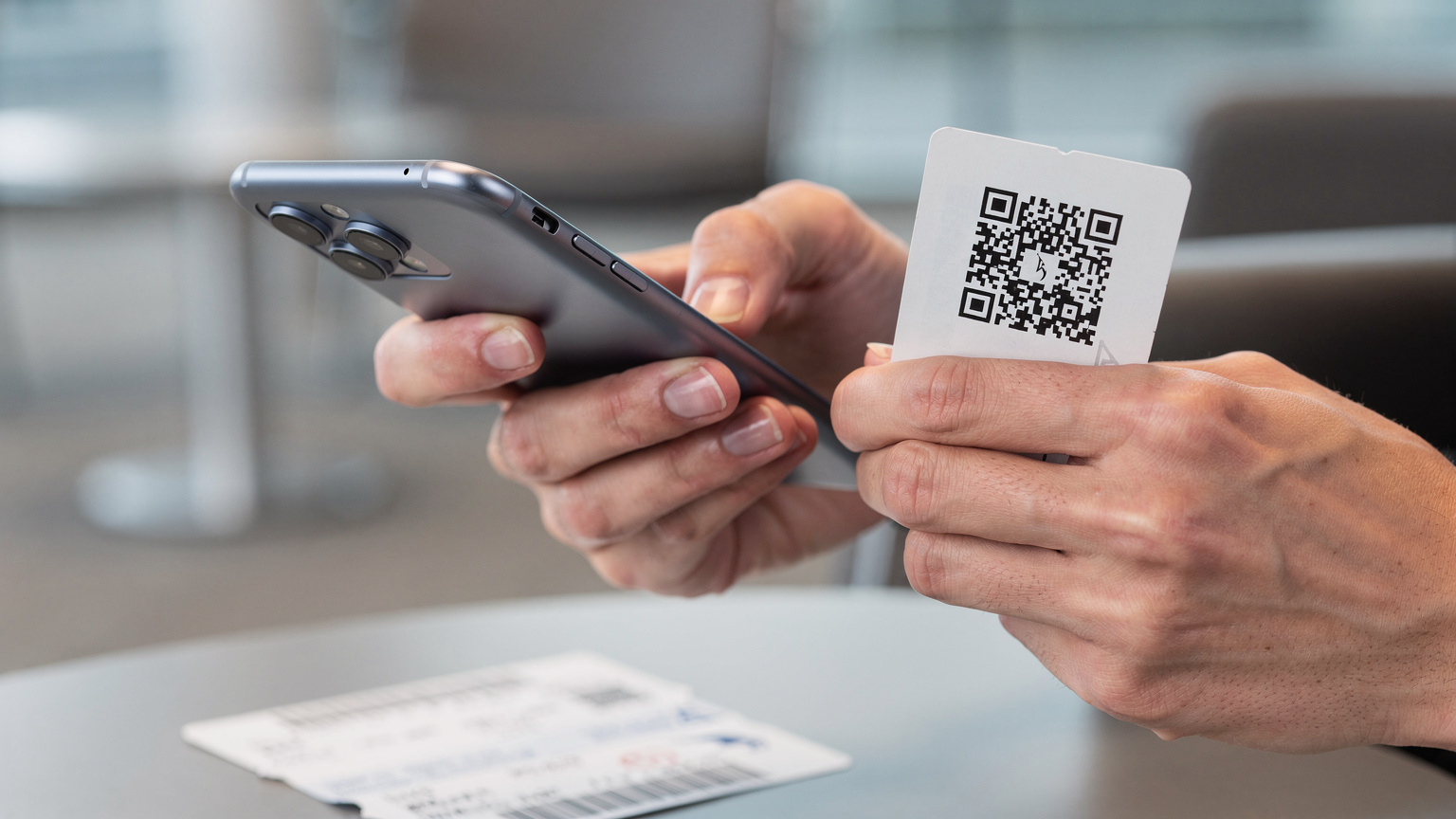 A close-up of a traveler's hands, in a well-lit airport lounge, carefully scanning an eSIM QR code from a printed paper with their smartphone, while a boarding pass rests nearby.