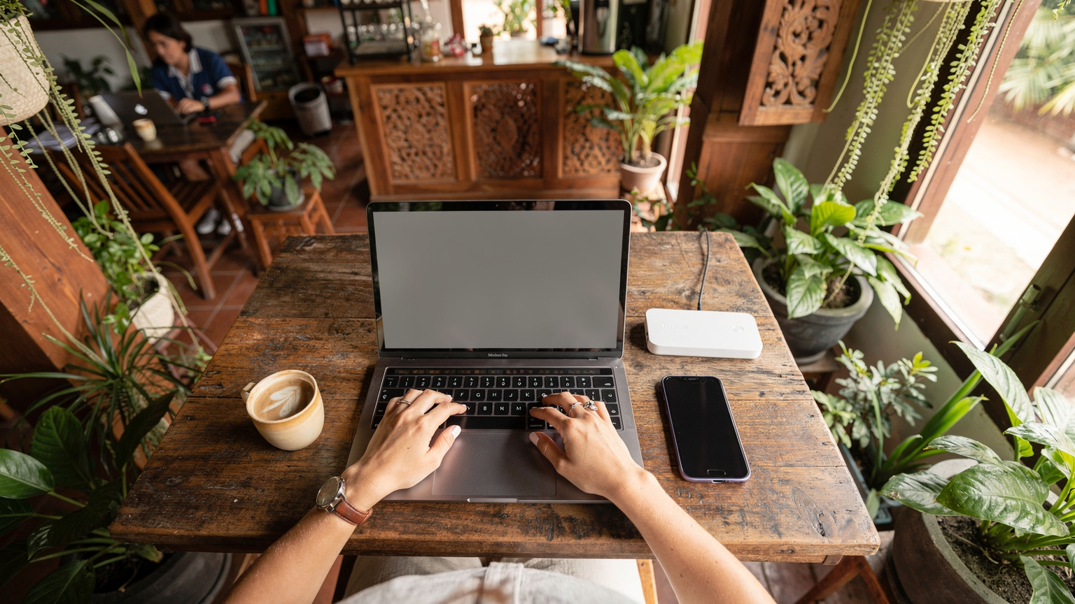 En travel digital nomade jobber fra en laptop på en kafé i Siem Reap, Kambodsja, med telefonen ved siden av som en hotspot. Kafeen er full av planter og har en avslappet atmosfære.