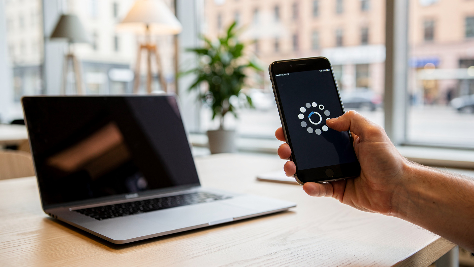 En person holder en smarttelefon med en aktiv hotspot-indikator, mens en laptop ligger åpen i forgrunnen, klar til å koble seg til. Bakgrunnen viser et interiør av et moderne co-working space.