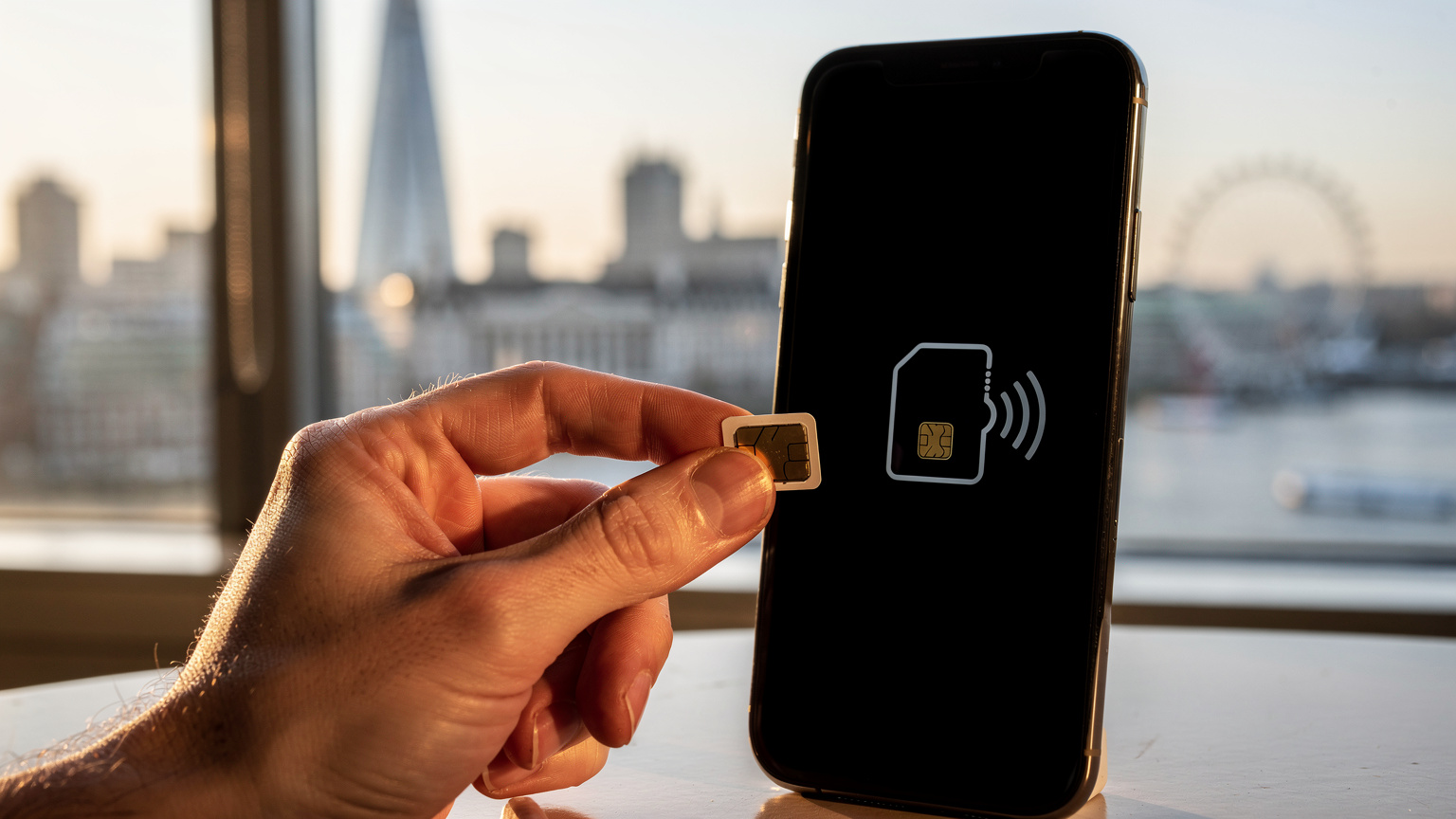 Primo piano di una mano che tiene una SIM fisica accanto a uno smartphone con un'icona eSIM sullo schermo, con lo skyline di Londra sfocato sullo sfondo.
