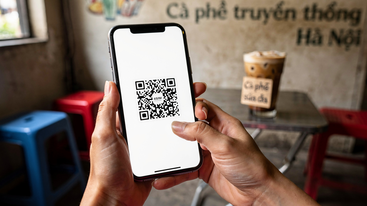 Cận cảnh bàn tay của một phụ nữ đang cầm iPhone 11, màn hình hiển thị mã QR để quét eSIM. Phía sau là khung cảnh một quán cà phê truyền thống ở Hà Nội với những chiếc ghế nhựa và ly cà phê sữa đá, tạo cảm giác thân thuộc.