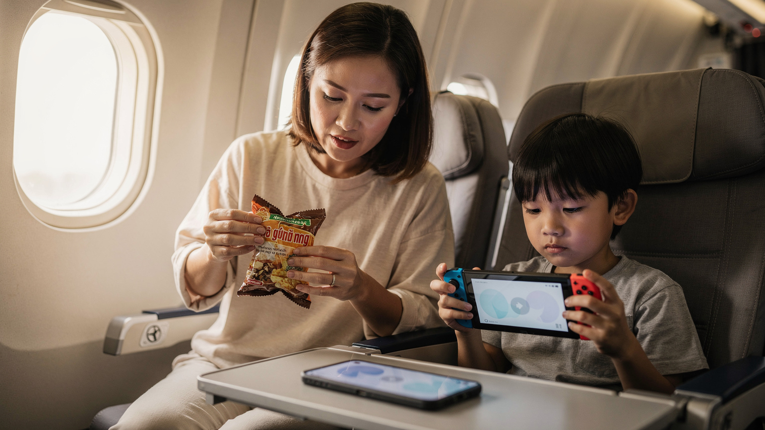 Một người phụ nữ Việt Nam đang ngồi bên cửa sổ máy bay, mở gói snack Việt Nam cho con trai nhỏ, người đang chơi game trên Nintendo Switch. Phía trước họ là một chiếc iPhone 11 đang hiển thị màn hình cài đặt mạng.