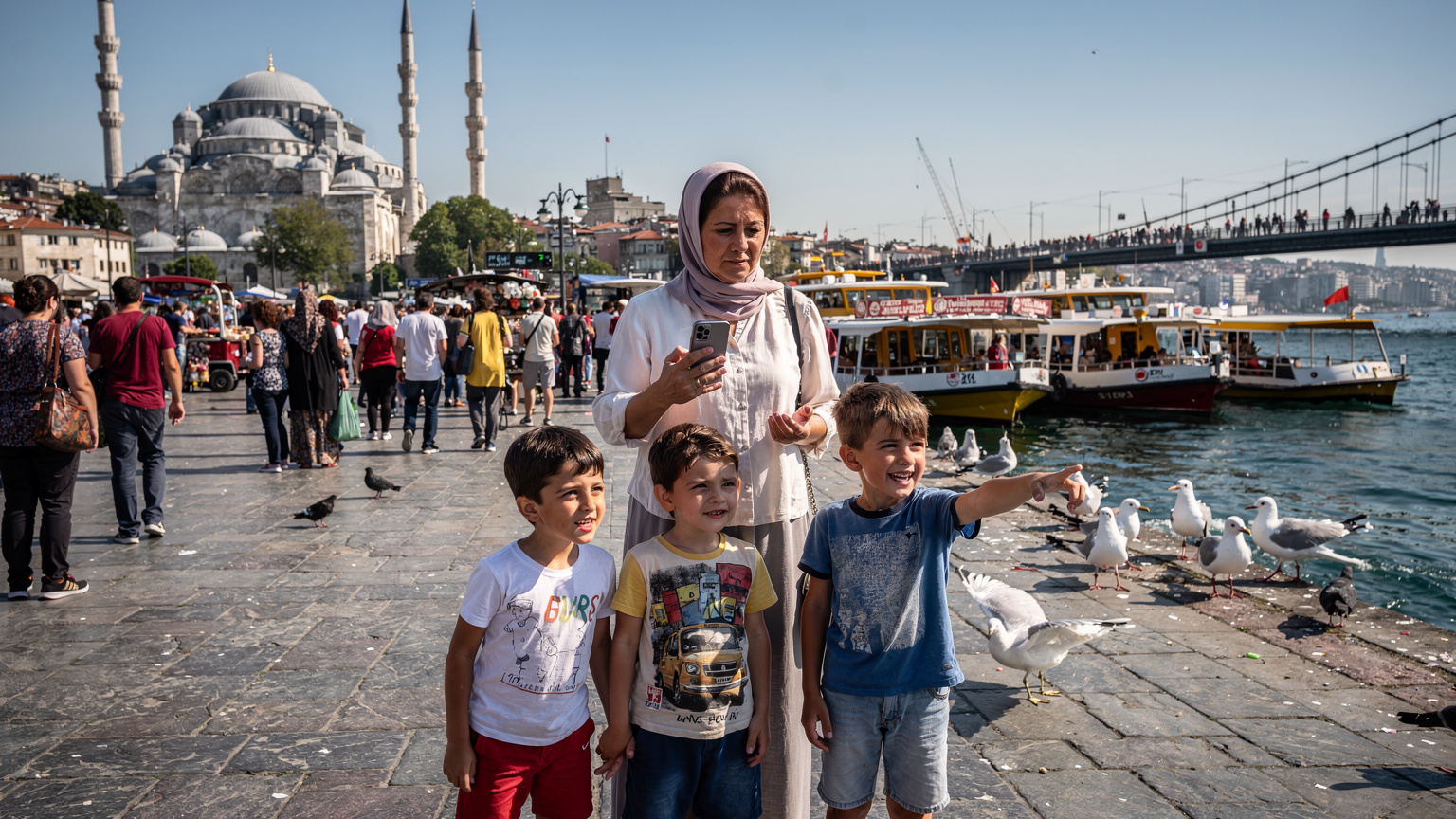 İstanbul'un Eminönü İskelesi'nde, elinde akıllı telefonuyla feribot saatlerini kontrol eden bir anne, yanında heyecanla martıları izleyen iki çocuğu. Arka planda Yeni Cami ve Galata Köprüsü.