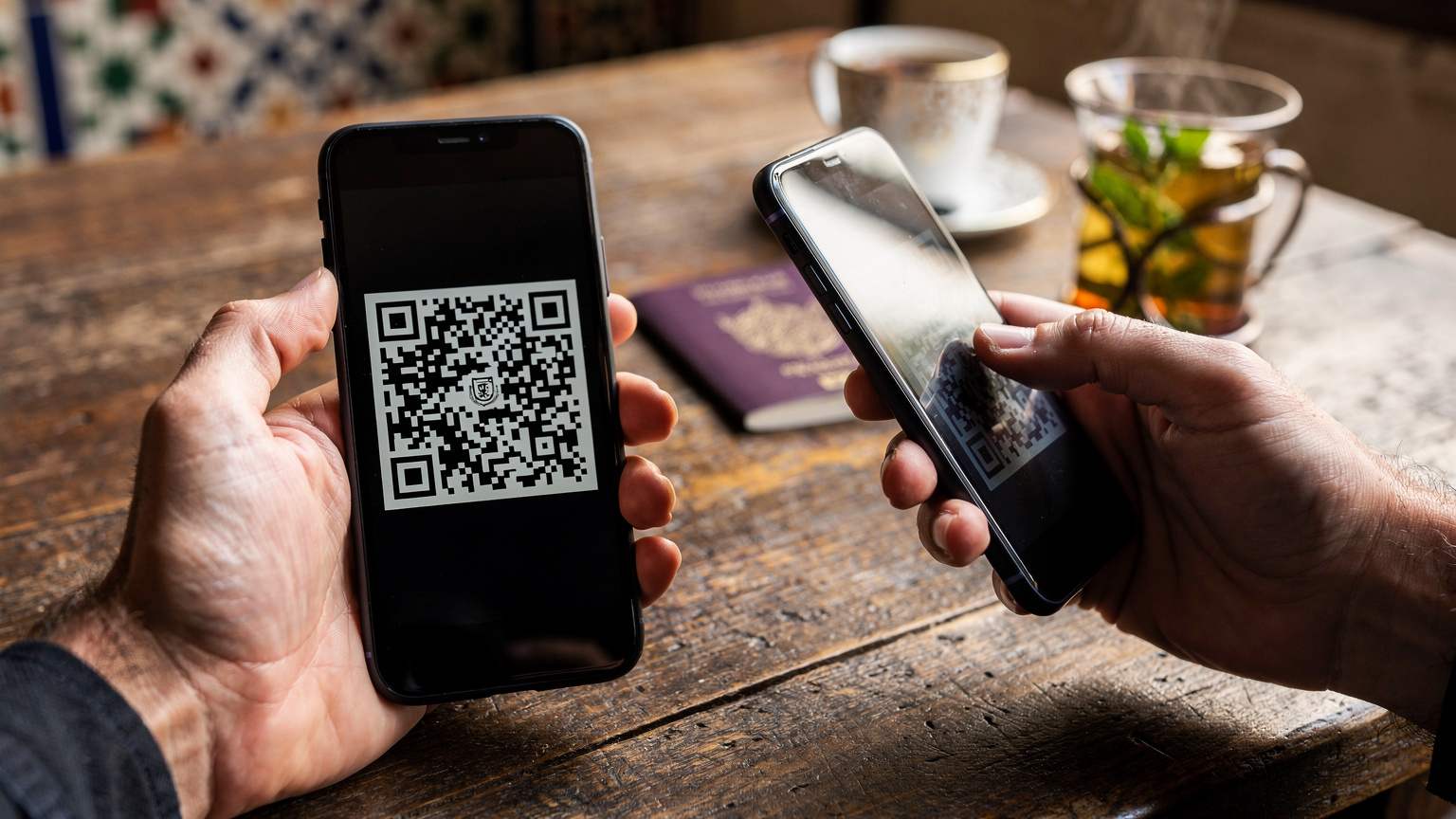 Un gros plan sur des mains utilisant un smartphone pour scanner un QR code, avec un passeport français et une tasse de thé à la menthe marocain en arrière-plan, sur une table en bois d'un café à Rabat.