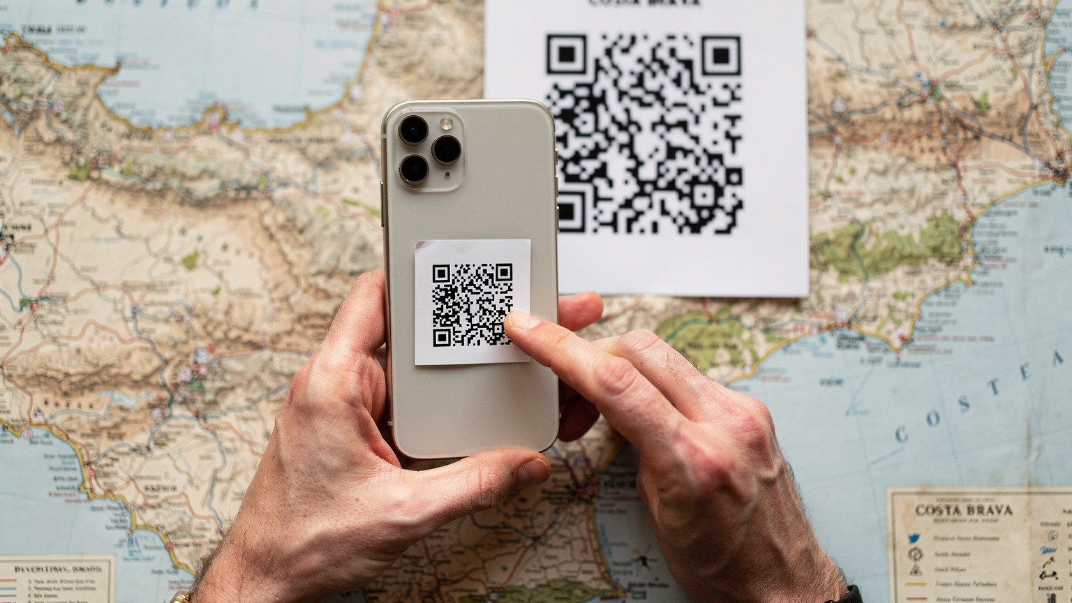 Manos de una persona usando su iPhone para escanear un código QR de activación de eSIM, con un mapa de la Costa Brava de fondo.