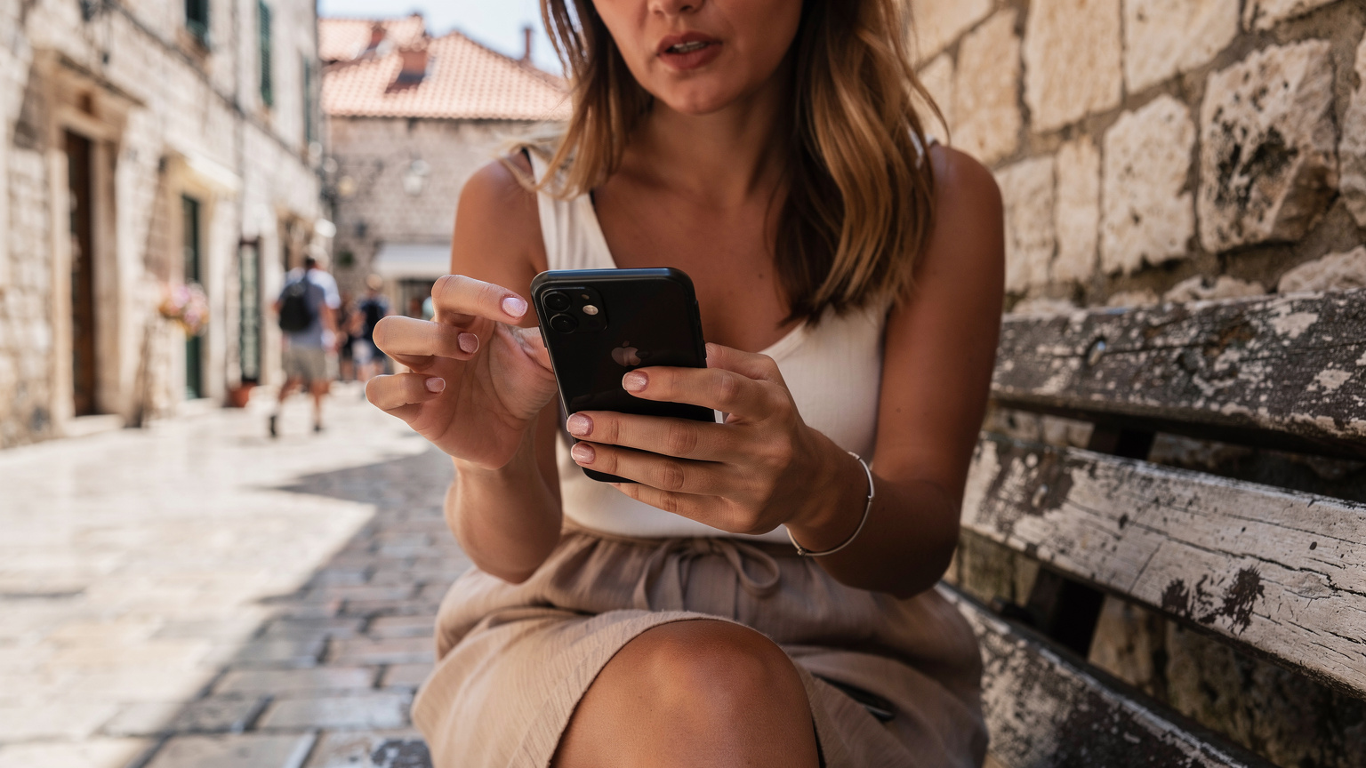 Zbliżenie na dłonie turystyki, która z lekkim zaniepokojeniem patrzy na ekran smartfona, próbując rozwiązać problem z połączeniem internetowym podczas podróży. W tle niewyraźne, ale rozpoznawalne uliczki Dubrownika.
