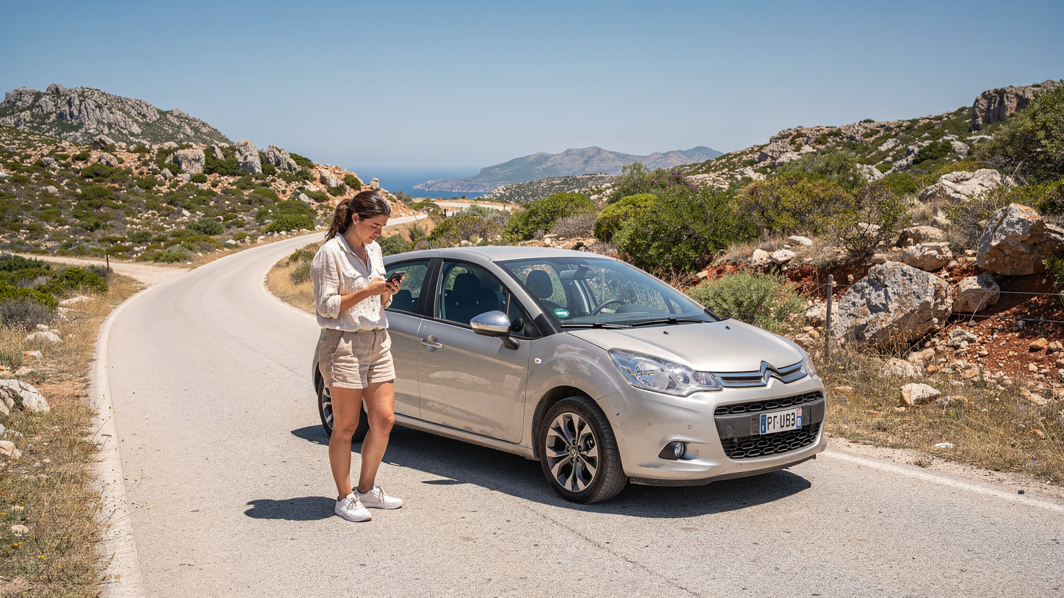 Eine junge Frau schaut besorgt auf ihr Smartphone, während sie auf einer verlassenen Landstraße in Sardinien steht, im Hintergrund ein Mietwagen und karge, steinige Landschaft unter einem strahlend blauen Himmel.
