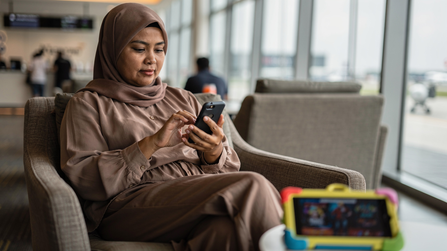 Seorang ibu sedang memuat turun aplikasi peta luar talian ke telefonnya di ruang menunggu lapangan terbang yang tenang, dengan tablet anak-anak di sisinya.
