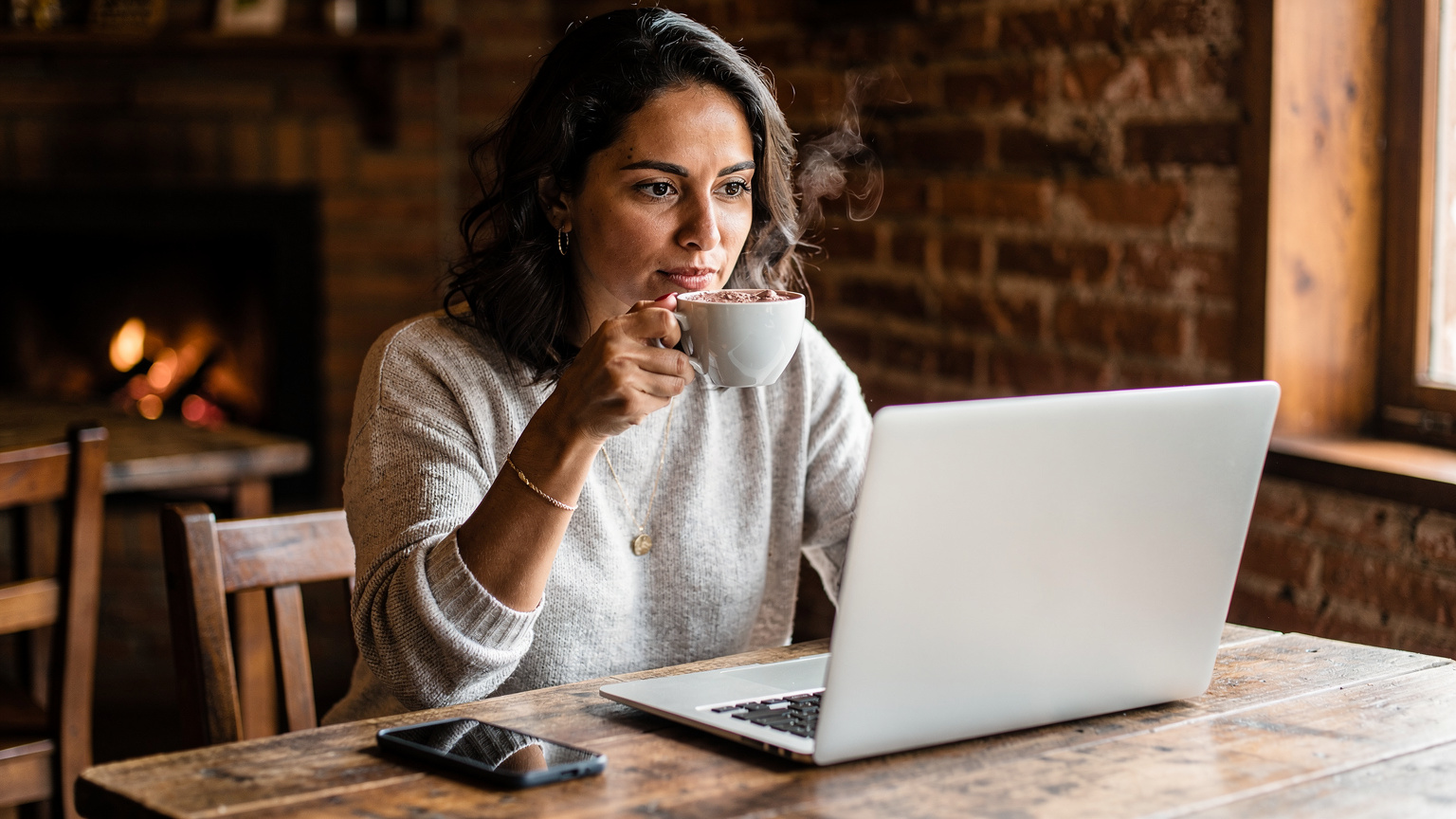 Uma mulher em um café aconchegante em Monte Verde, Minas Gerais, com a tela do laptop aberta e um smartphone ao lado, desfruta de um chocolate quente, usando a internet para planejar seu dia.