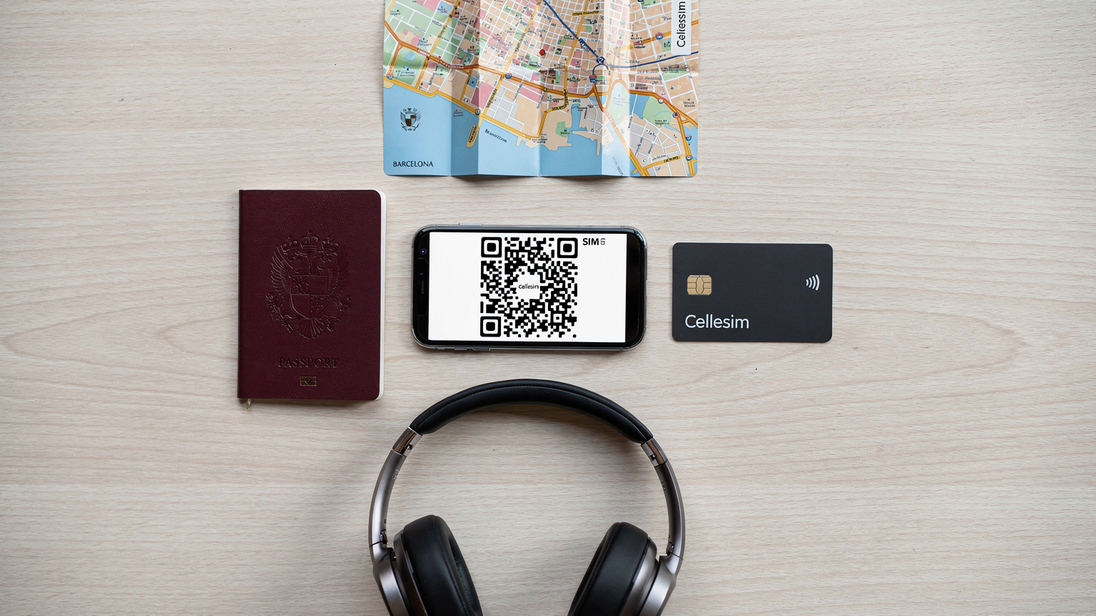 An editorial overhead flat-lay of travel essentials: a passport, a smartphone displaying a Cellesim eSIM QR code, a Cellesim branded payment card, noise-canceling headphones, and a small, folded map of Barcelona.