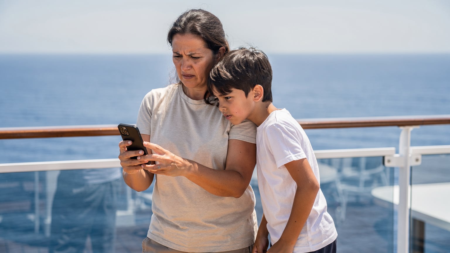 A concerned parent and their child on a cruise ship deck, both looking intently at a smartphone screen, depicting the common frustration with slow or expensive onboard internet.