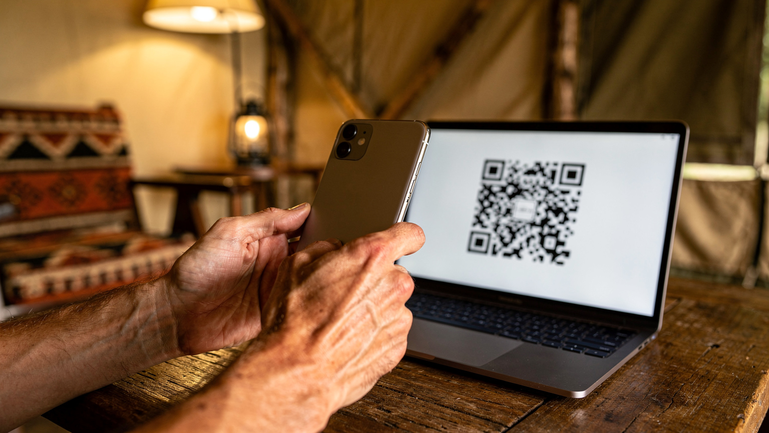Zbliżenie na dłonie osoby instalującej eSIM na smartfonie, z niewyraźnym tłem wnętrza obozu safari i widocznym kodem QR na ekranie laptopa.