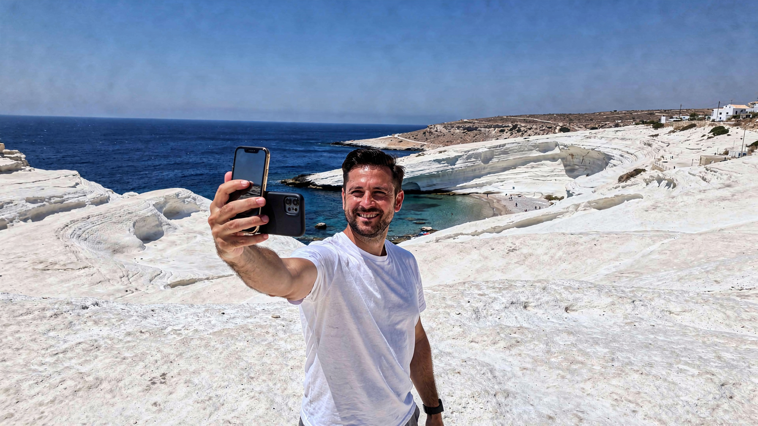 Un homme, l'air serein, tient son smartphone à bout de bras pour une photo, avec en arrière-plan la plage lunaire de Sarakiniko à Milos, Grèce, et les formations rocheuses blanches caractéristiques.