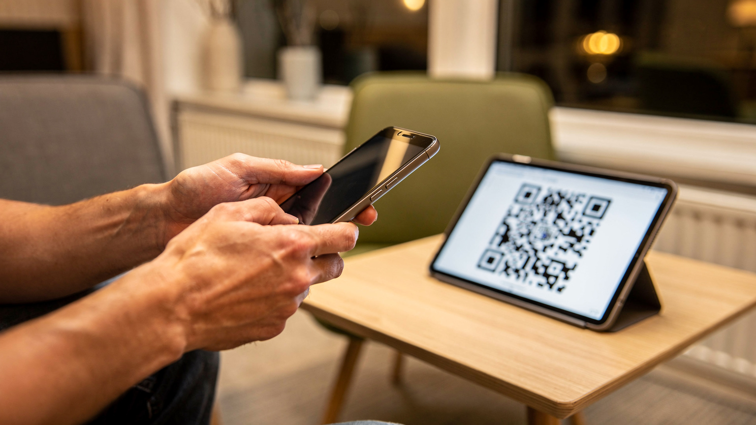 En persons hånd holder en smartphone, der er klar til at scanne en QR-kode. QR-koden vises på en tabletskærm i baggrunden, placeret på et lille bord i en hyggelig, moderne hotelobby i Grønland med elementer af nordisk design.