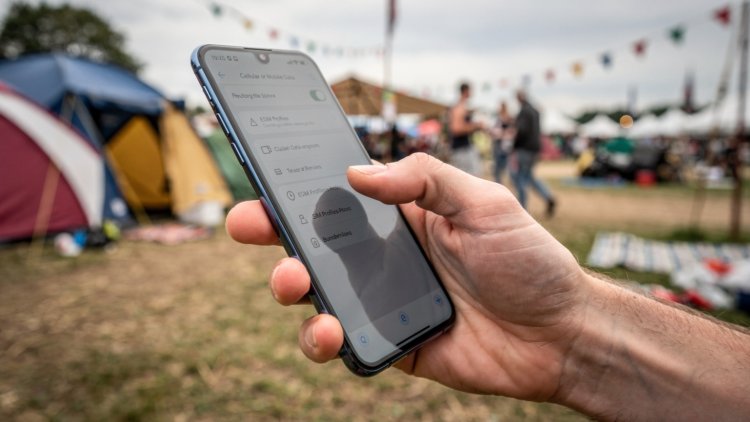 En närbild på en hand som håller en smartphone och bläddrar genom eSIM-inställningar, med en suddig bakgrund av en festivalcamping.