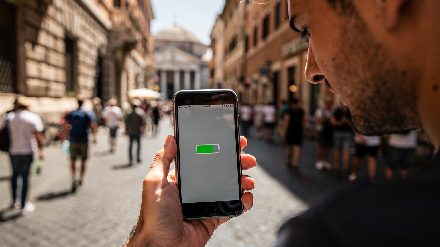 Una mano tiene in mano un telefono mostrando una schermata di errore o di batteria scarica, con uno sfondo sfocato di una strada affollata di Roma, suggerendo un momento di frustrazione tecnologica.