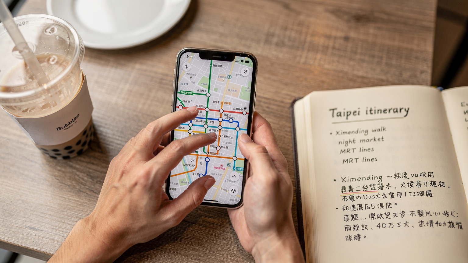 Cận cảnh một bàn tay đang thao tác trên màn hình điện thoại thông minh, hiển thị ứng dụng bản đồ với tuyến đường MRT ở Đài Loan, với tách trà sữa trân châu và quyển sổ tay du lịch trên bàn, gợi cảm giác lập kế hoạch và khám phá.