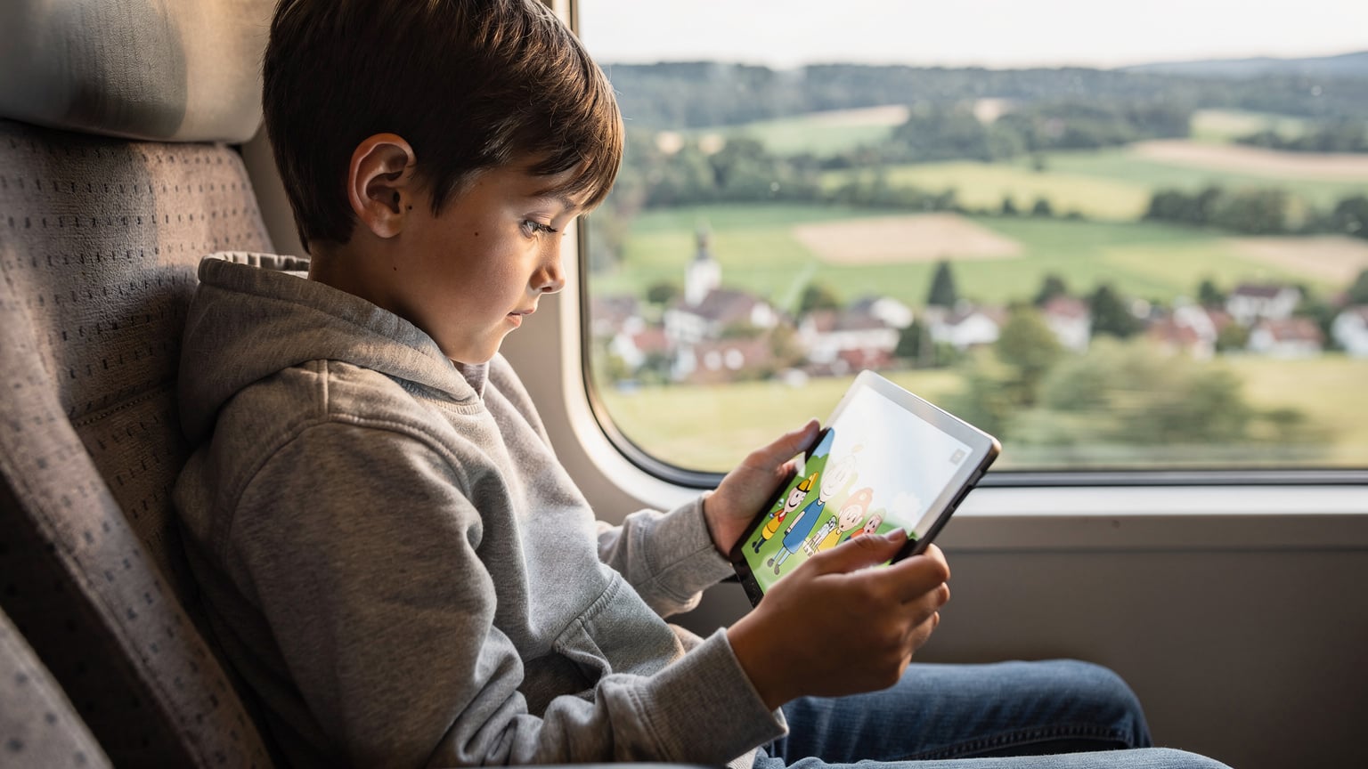 A child, around 10 years old, intently focused on a tablet displaying a cartoon, while sitting comfortably in a train seat with a scenic European countryside blurring past the window.