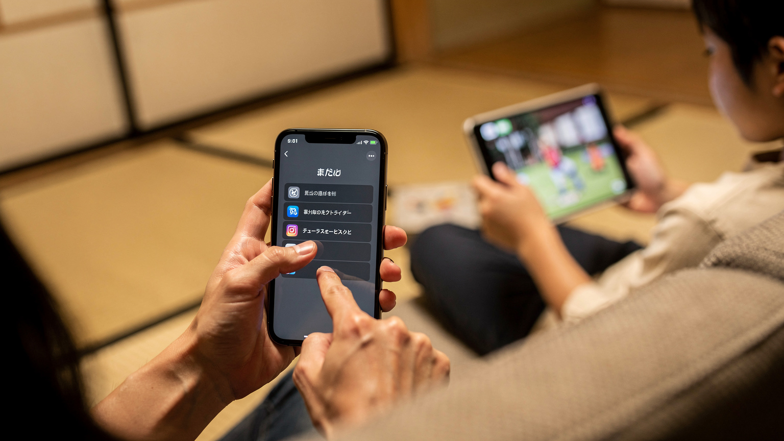 子供が遊んでいるiPadに、親がスマートフォンからスクリーンタイム設定を行っている手元のクローズアップ。