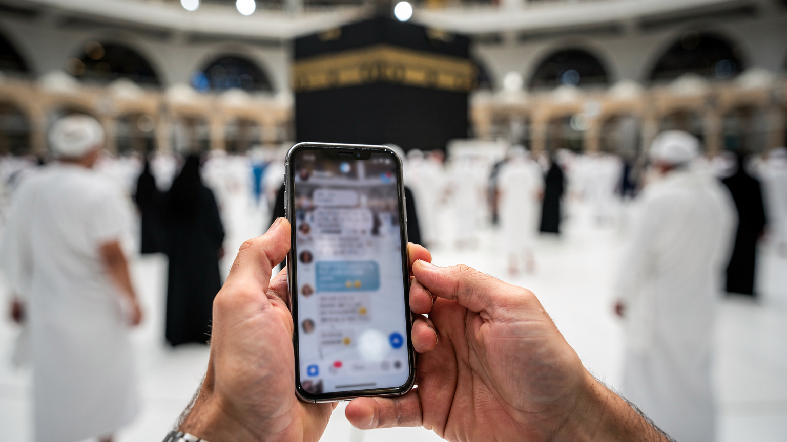 Tangan seorang jemaah haji memegang ponsel di tengah keramaian jamaah lain yang sedang tawaf di Masjidil Haram, dengan notifikasi aplikasi chat keluarga terlihat samar di layar, menggambarkan koneksi di tengah spiritualitas.
