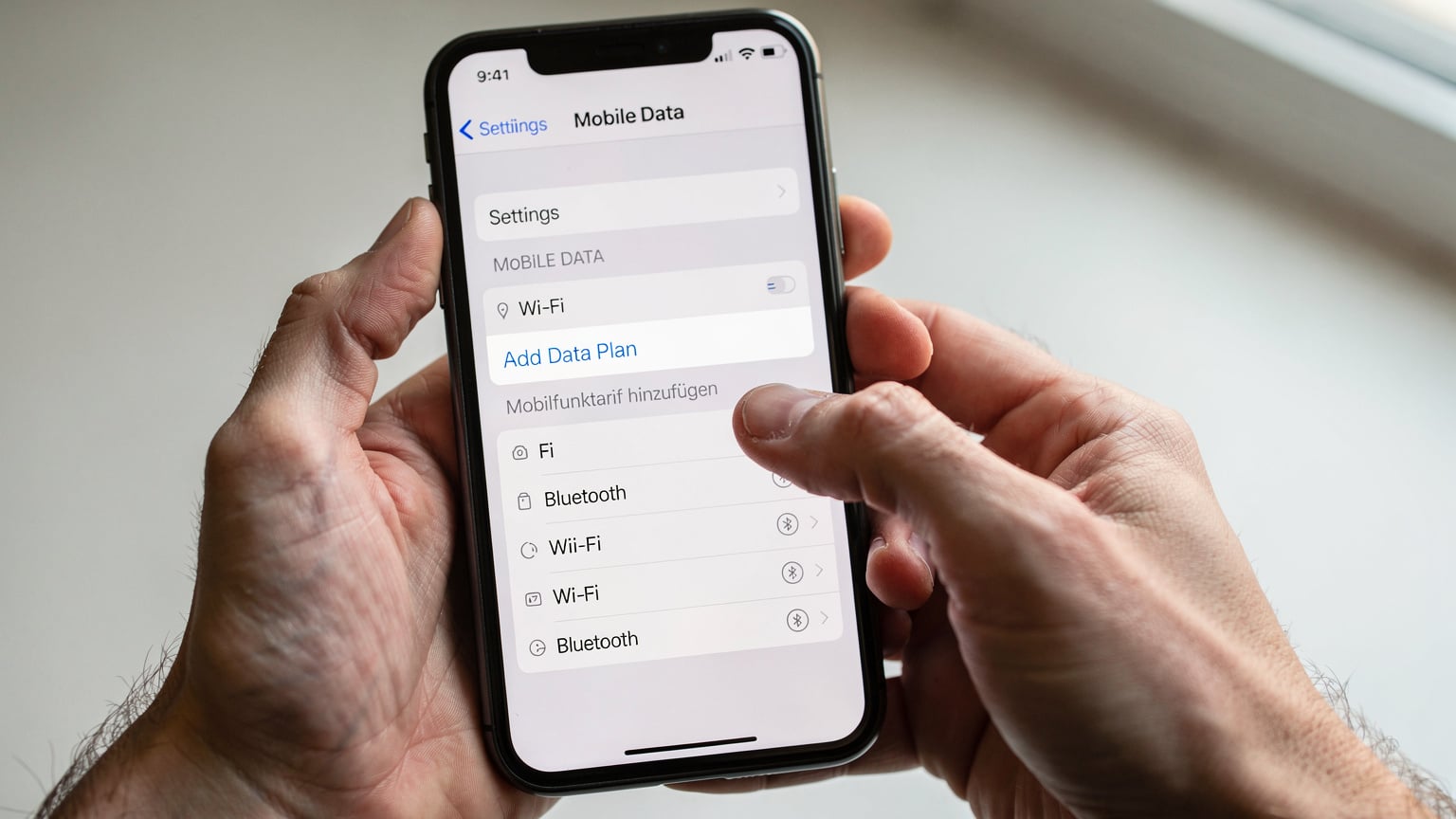 Eine Nahaufnahme zeigt Hände, die ein iPhone halten und die Einstellungen für Mobilfunkverbindungen überprüfen, mit einem Fokus auf die Option 'Mobilfunktarif hinzufügen'.
