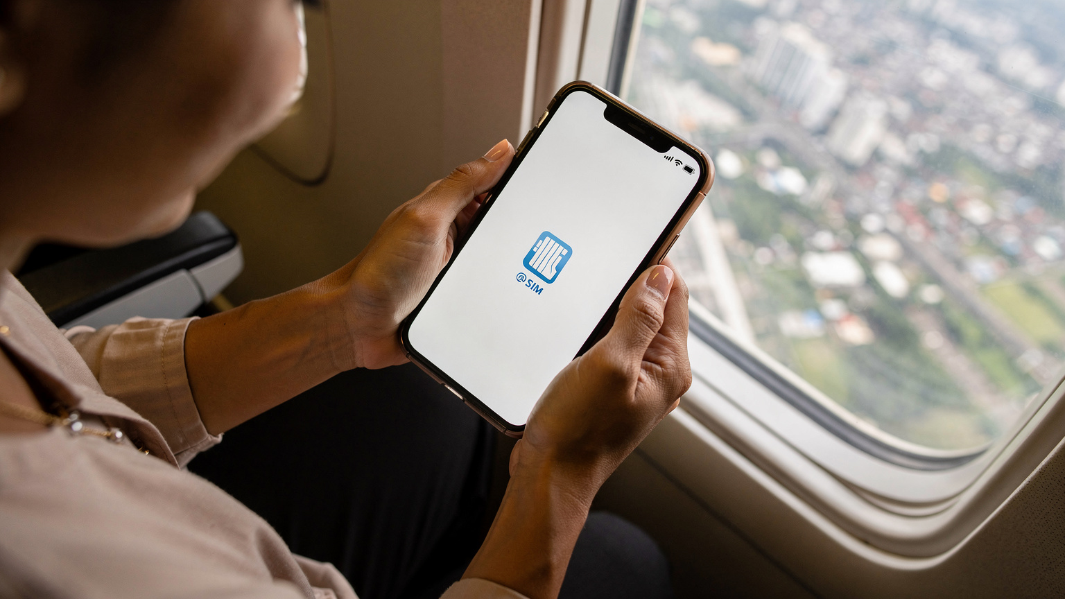 Tangan seorang ibu memegang smartphone yang menampilkan notifikasi 'eSIM aktif' dengan sinyal penuh, tersenyum lega di depan jendela pesawat yang memperlihatkan pemandangan kota di bawah.