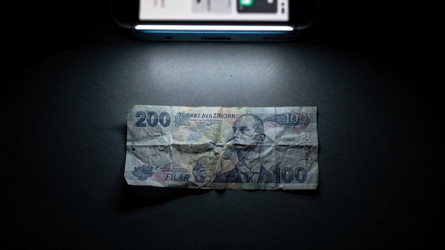 Bir Türk Lirası banknotunun üzerinde, yurt dışı seyahat faturasını gösteren bir telefon ekranının yansıması, roaming ücretlerinin maliyetini sembolize ediyor.
