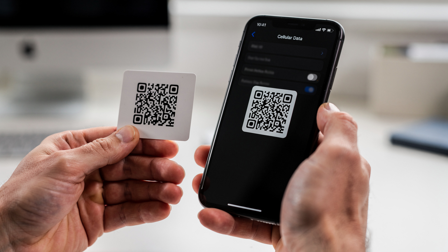 Une main tient un iPhone affichant un écran de réglages de données cellulaires, tandis qu'une autre main tend une petite carte avec un QR code imprimé, prête à être scannée pour l'activation d'une eSIM.