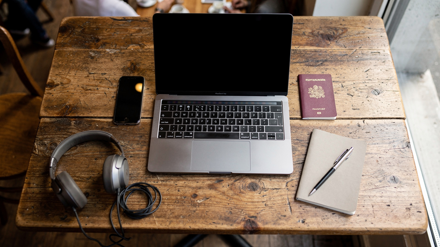 Une vue aérienne d'une table de café avec un ordinateur portable ouvert, un iPhone, un passeport français et quelques notes éparses, illustrant l'organisation d'un nomade digital pour le travail et le voyage.