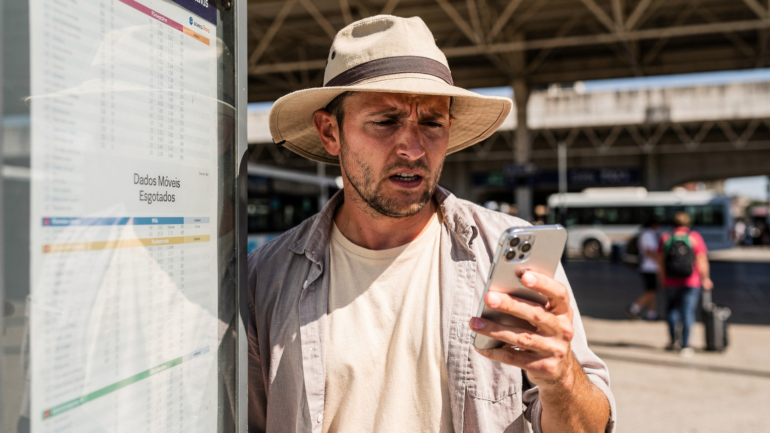 Um viajante com um chapéu de sol, olhando com preocupação para o ecrã do seu iPhone 18, que mostra uma notificação de dados móveis esgotados, num terminal de autocarros em Sete Rios, Lisboa.