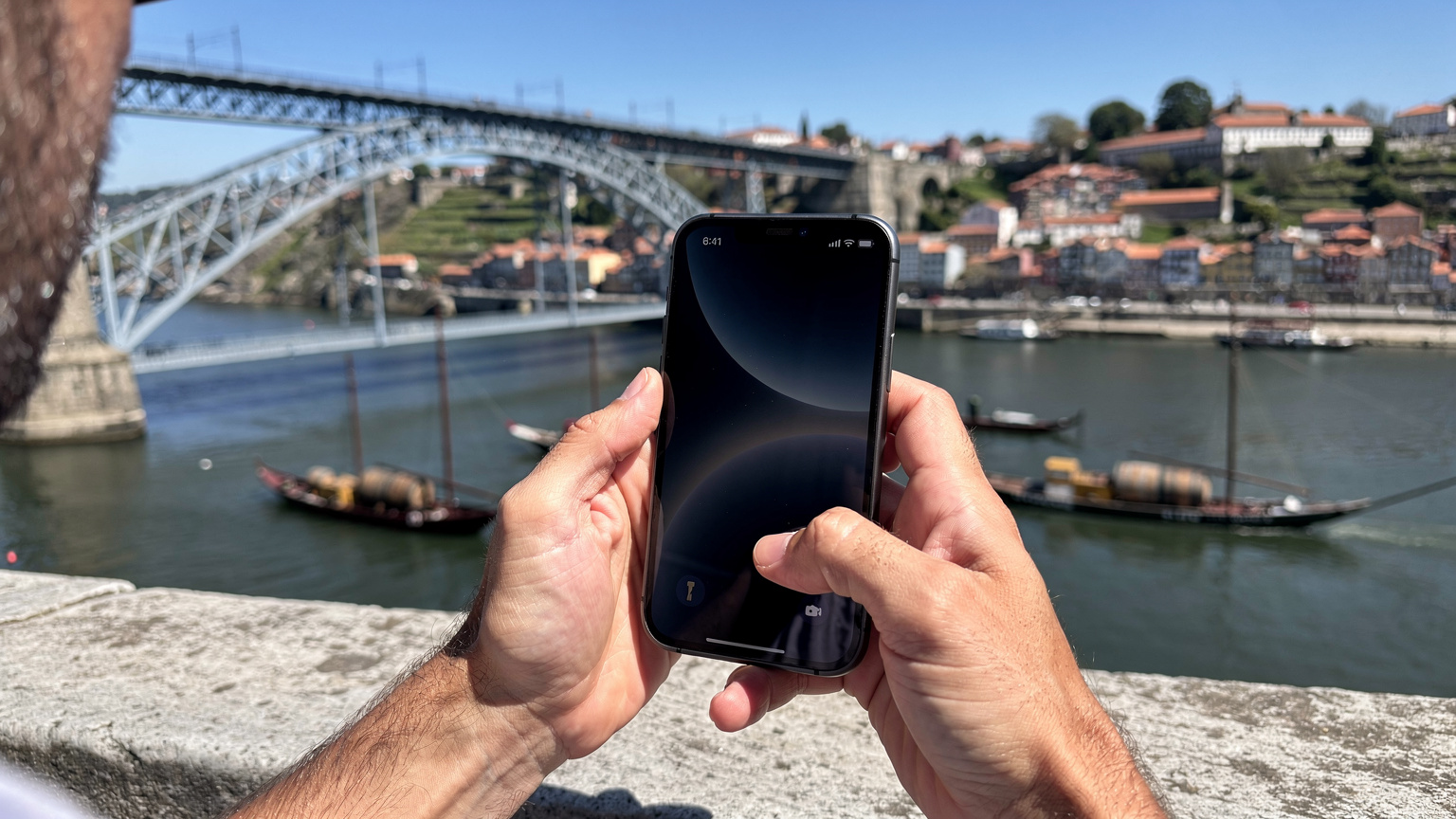 Mãos de um homem a segurar um iPhone 18, exibindo a tela de configurações de eSIM em frente à Ponte D. Luís I e ao rio Douro, no Porto, durante um dia ensolarado.