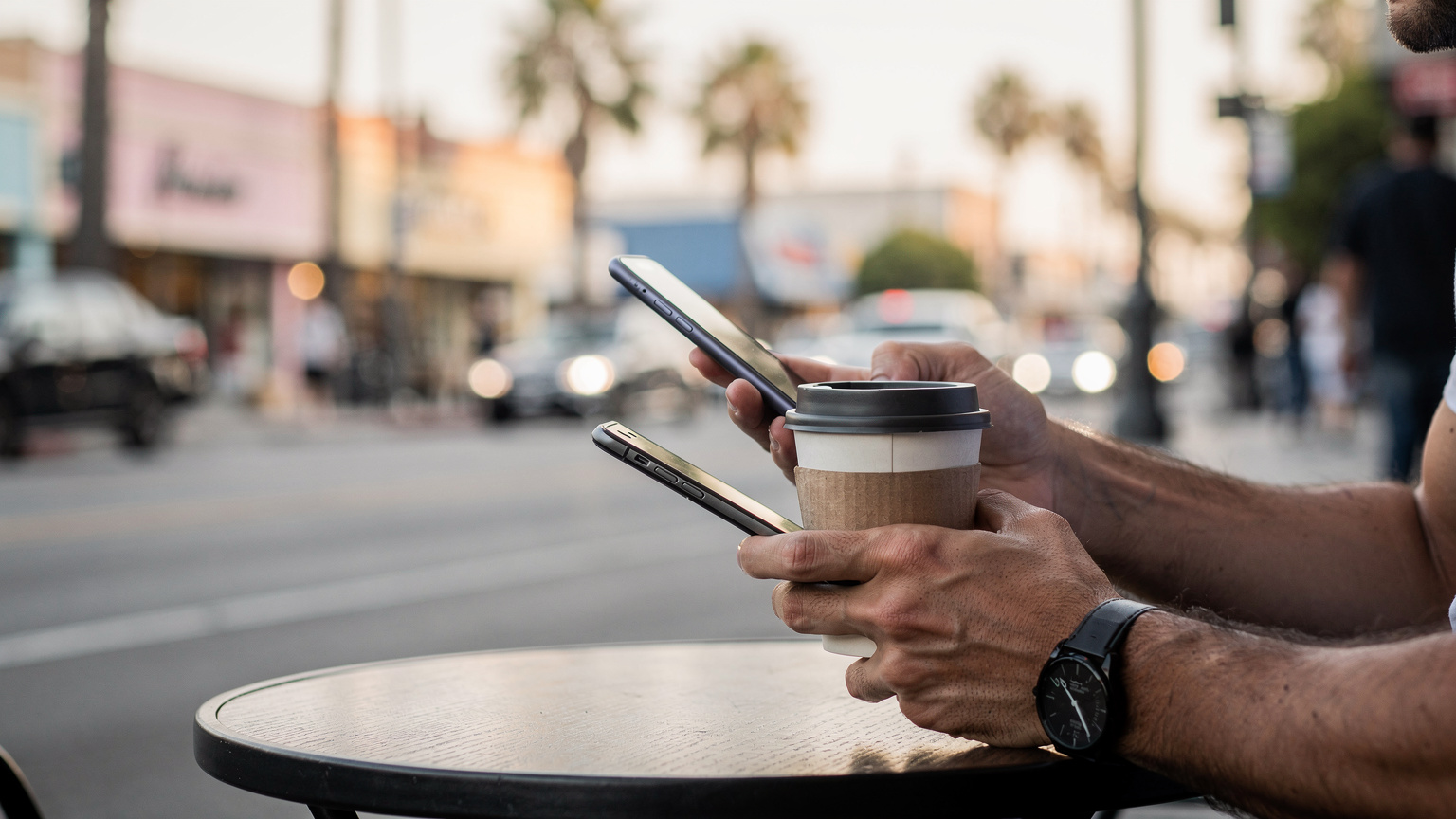 Un primo piano delle mani di una persona che tiene uno smartphone e una tazza di caffè, con una strada affollata di Los Angeles sullo sfondo, suggerendo una pausa in un caffè all'aperto.
