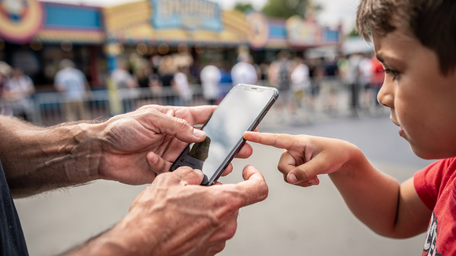 Manos de un padre y un niño interactuando con un teléfono, ajustando la configuración de datos mientras esperan en la fila para una atracción en un parque temático europeo.
