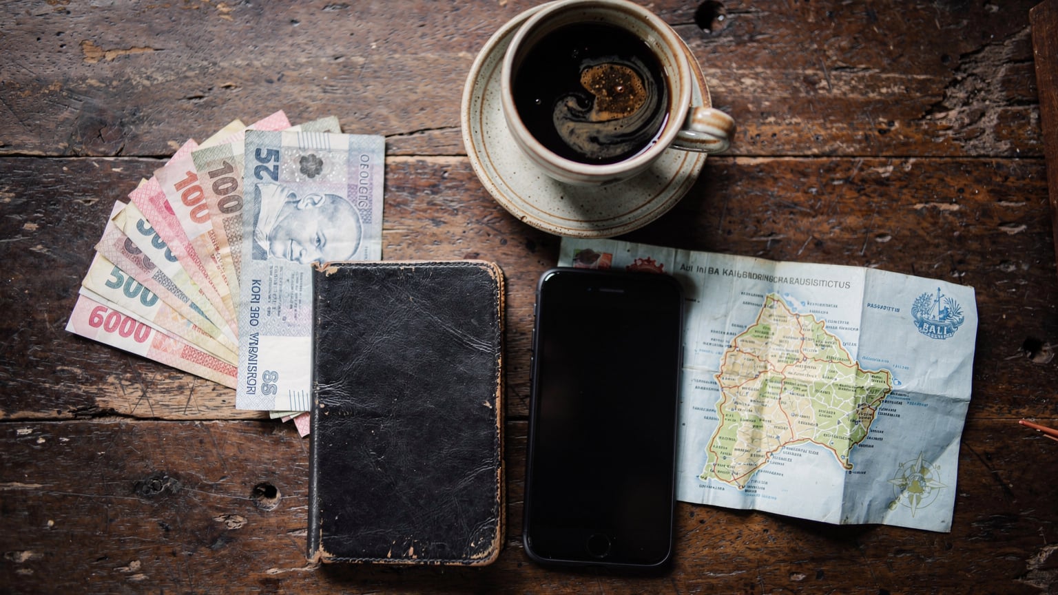 An overhead flat-lay composition of a backpacker's travel essentials: a worn passport, a smartphone, a small wad of Indonesian rupiah, a folded local map, and a traditional cup of Indonesian kopi tubruk, all arranged on a rustic wooden table.