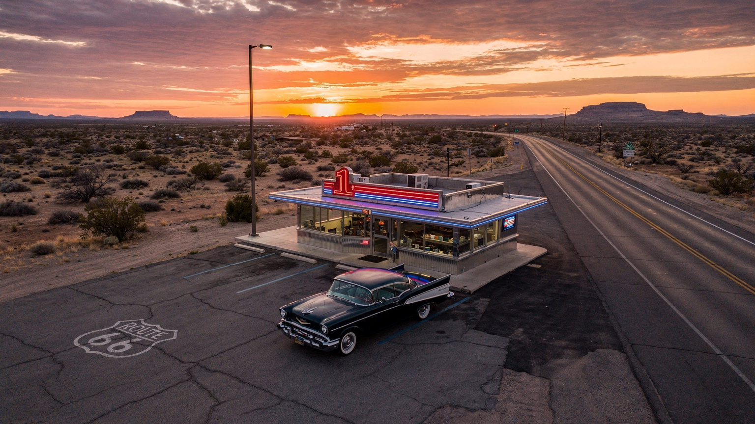 Una veduta aerea di un classico diner americano su un tratto isolato della Route 66, con un'auto d'epoca parcheggiata fuori e il sole che tramonta all'orizzonte, evocando un senso di avventura e libertà.