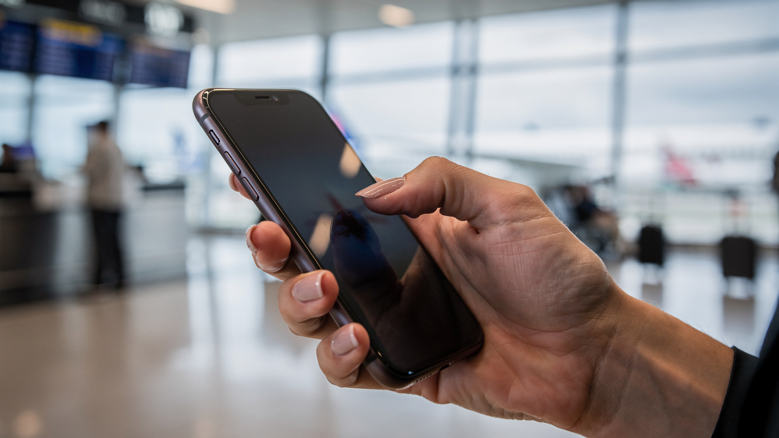 Uma mão segurando um smartphone, com os dedos digitando em uma tela embaçada, enquanto o reflexo de um terminal de aeroporto pode ser visto no plano de fundo, transmitindo uma sensação de urgência.