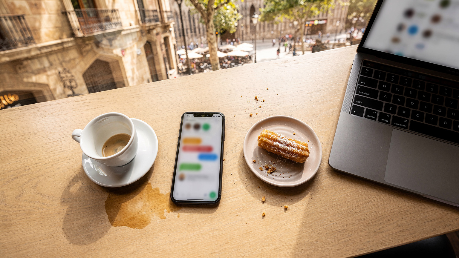 Uma mesa de café em Barcelona com um smartphone exibindo a interface de um aplicativo de mensagens, um copo de café e um notebook aberto, com as Ramblas ao fundo em um dia ensolarado.