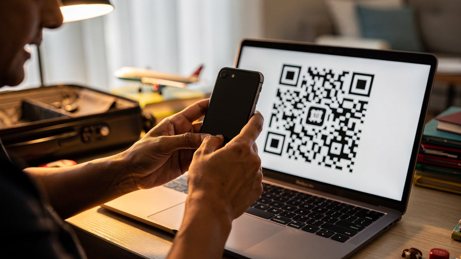 एक भारतीय यात्री अपने आईफोन पर Cellesim QR कोड को स्कैन कर रहा है, जो एक लैपटॉप की स्क्रीन पर दिखाया गया है, बैकग्राउंड में फैमिली ट्रैवल की तैयारी दिख रही है।