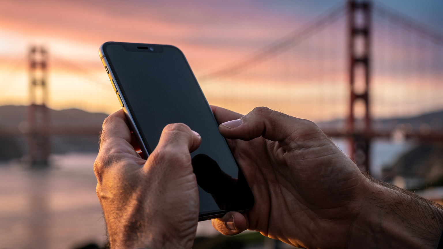 Een close-up van handen die een smartphone vasthouden, waarbij de persoon door de Cellesim-app scrollt om eSIM-dataplannen te vergelijken, met op de achtergrond een onscherp beeld van de Golden Gate Bridge bij zonsondergang.