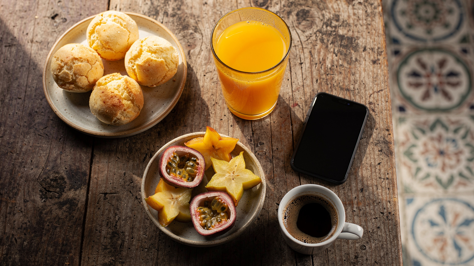 Uma vista aérea de uma mesa de café da manhã em um hotel boutique no Nordeste brasileiro, com um smartphone ao lado de um copo de suco de fruta tropical, pães frescos e frutas, simbolizando conectividade e relaxamento.