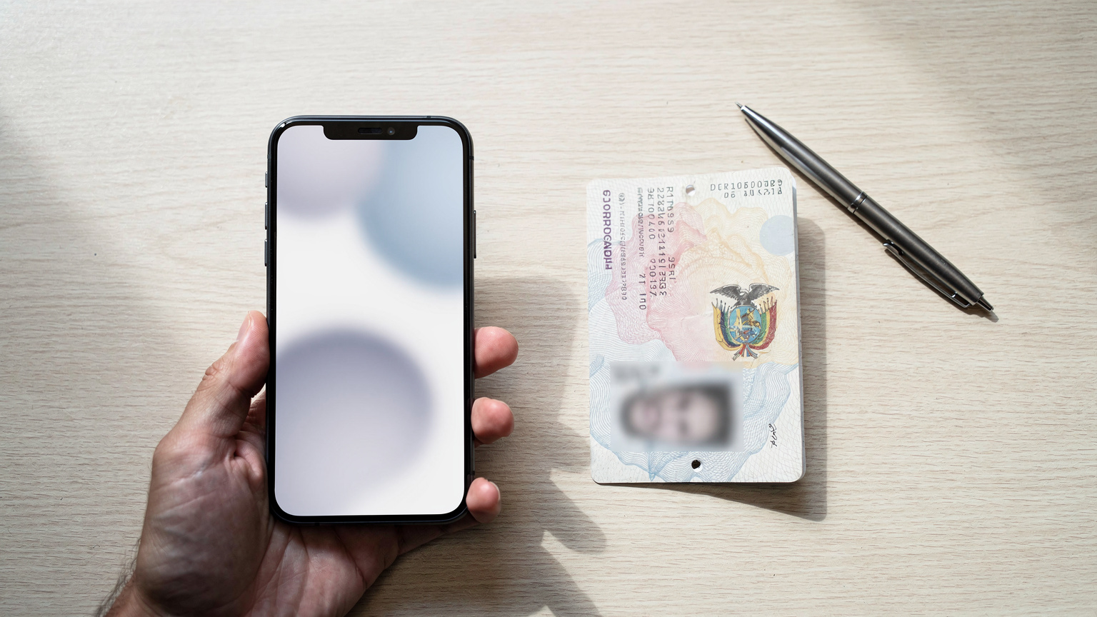 Une main tenant un smartphone affichant un formulaire administratif en ligne, avec un passeport équatorien ouvert et un stylo posés sur une table en bois clair à côté, le tout éclairé par la lumière naturelle d'une fenêtre.